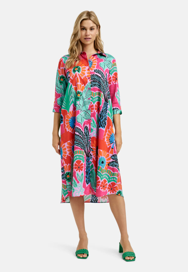Eine Frau in einem farbenfrohen Midikleid mit Blumenmuster steht mit neutralem Gesichtsausdruck da. Das Kleid ist in Rosa, Rot, Grün und Schwarz gehalten. Sie trägt grüne Schuhe. Der Hintergrund ist schlicht weiß.